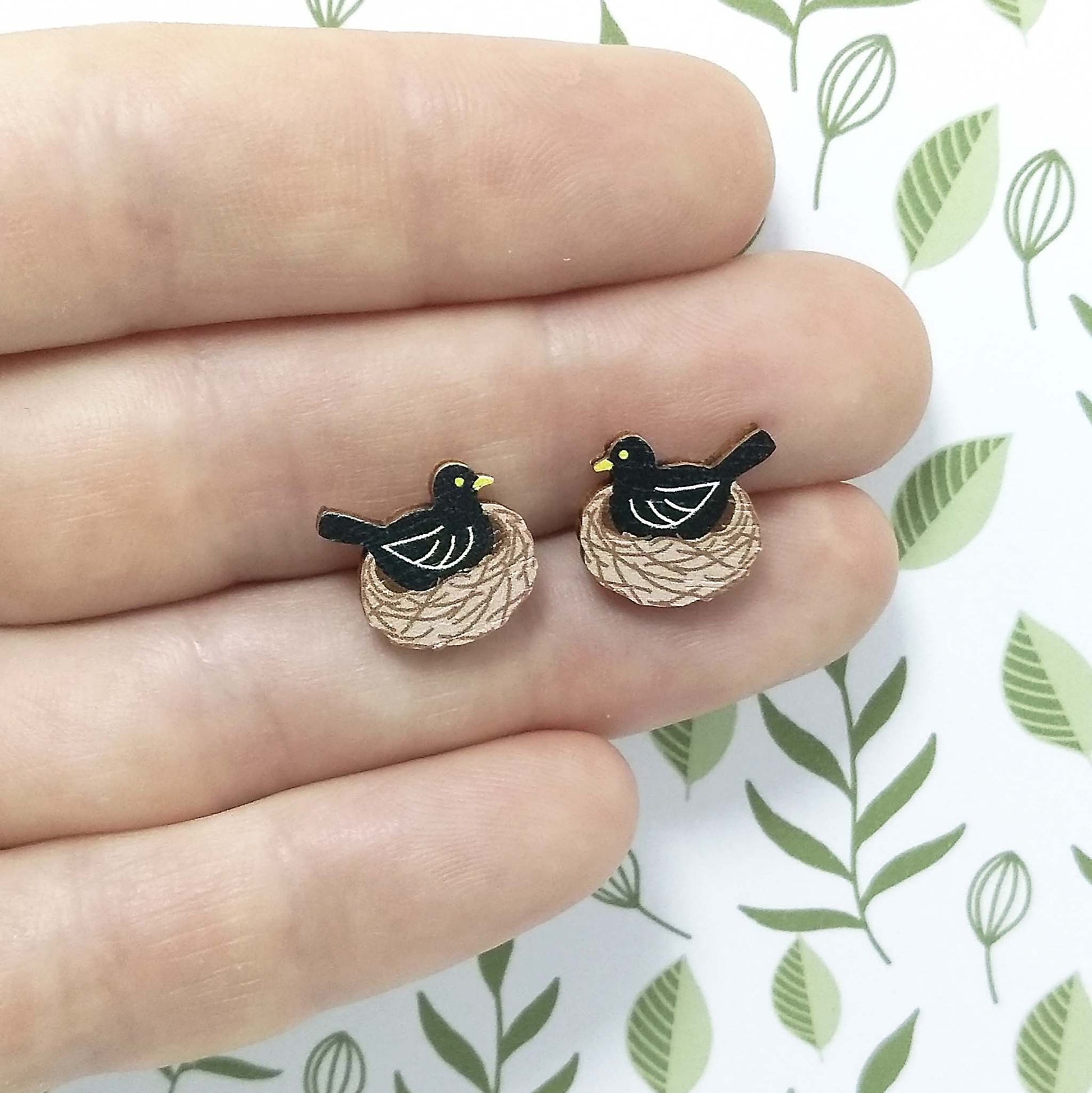 Wooden blackbird stud earrings held on hand, blackbird is sitting in nest and colours of black, brown and yellow